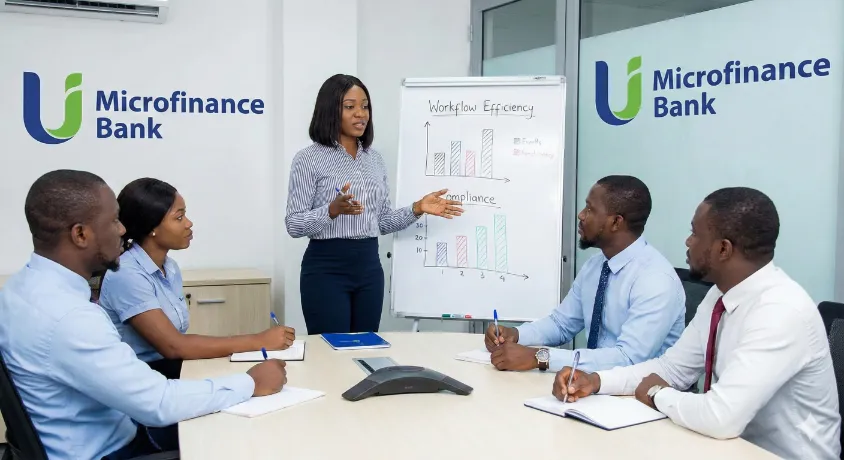 Female Operations Manager conducting a team meeting and training session regarding workflow efficiency and compliance strategies using a whiteboard in a U Microfinance Bank conference room.